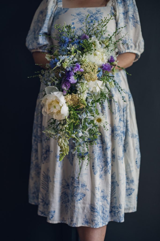 woman in blue and white china dress holding a waterfall bouquet of Cress, queen anne's lace, delphinium, white peony, white cosmo, purple sweet pea, nigella, yellow yarrow, lupine