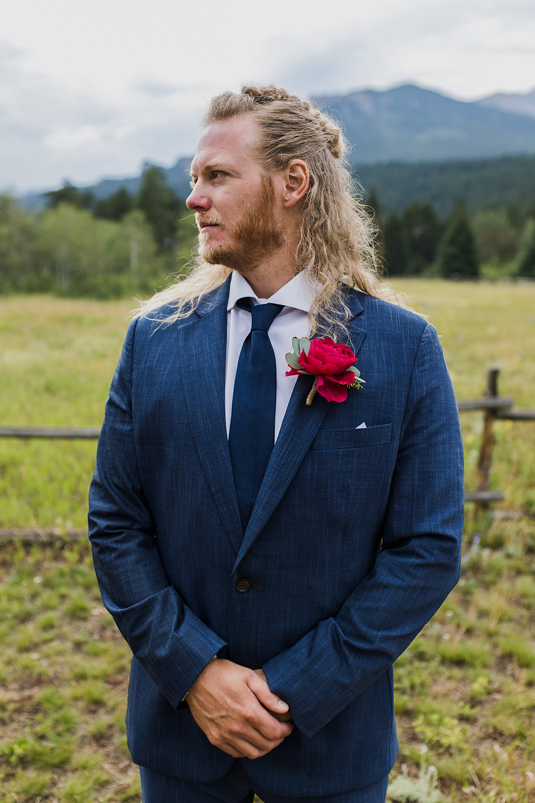 groom standing in a field wearing a blue suit with red rose boutonniere in estes park colorado wedding